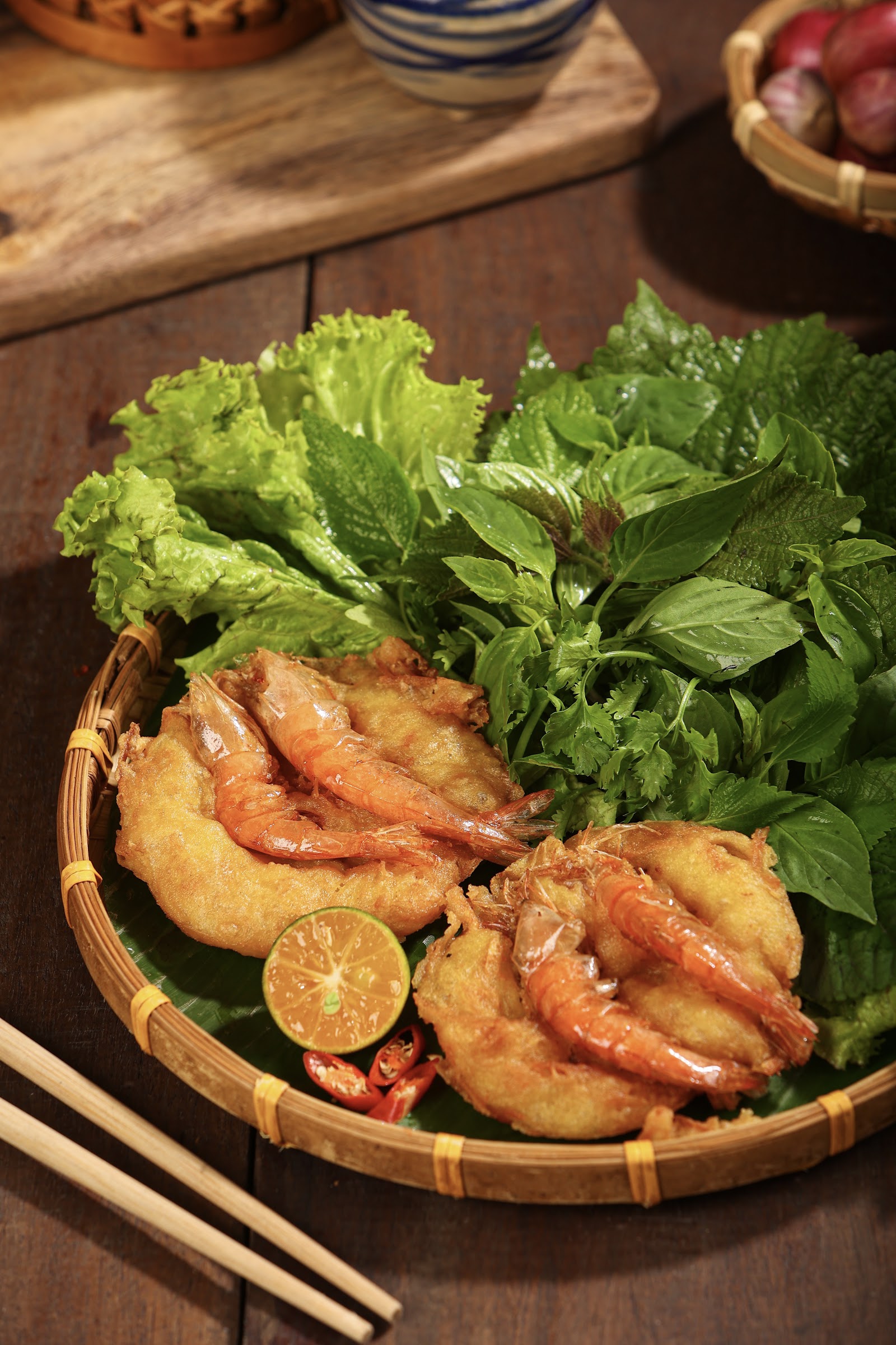 Two bánh tôm Hồ Tây with whole shrimp on banana leaf in a woven bamboo basket, with herbs, halved calamansi and chili