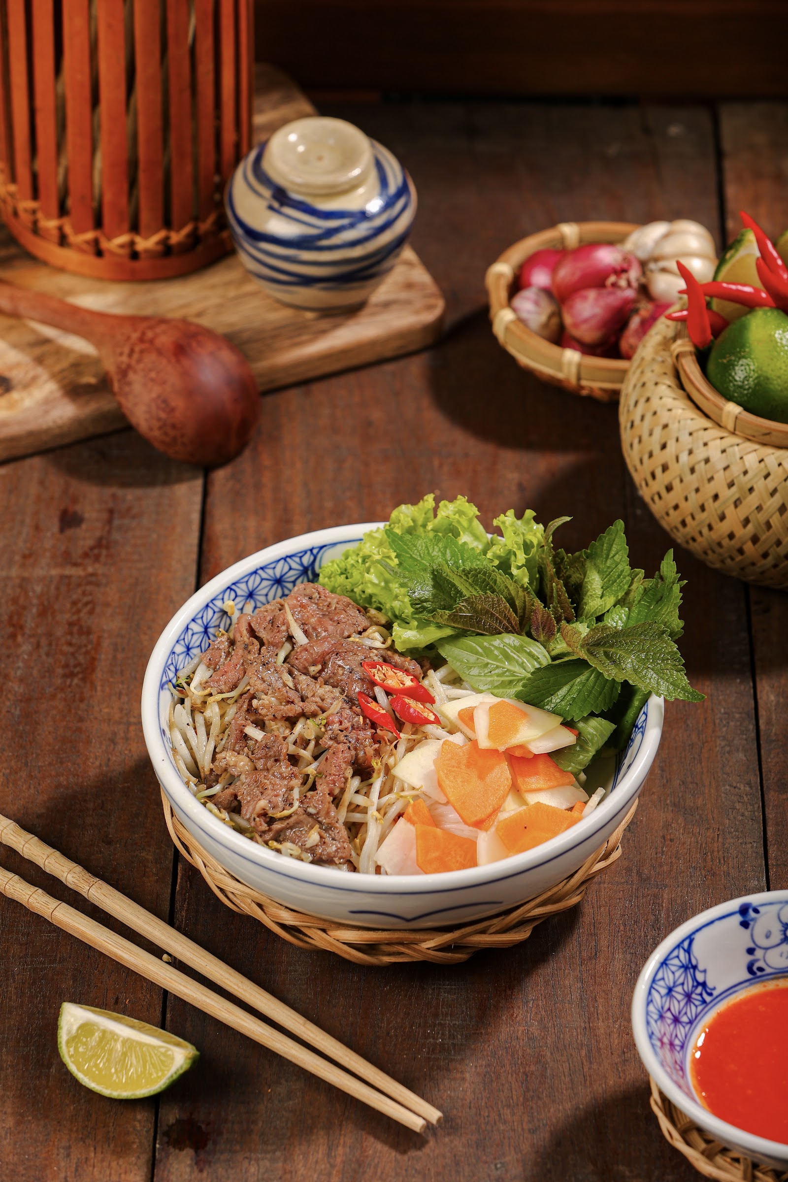 Bún bò Nam Bộ in a blue-white bowl on a woven tray: sautéed beef, vermicelli, bean sprouts, pickled carrot, herbs — lime and bamboo chopsticks beside