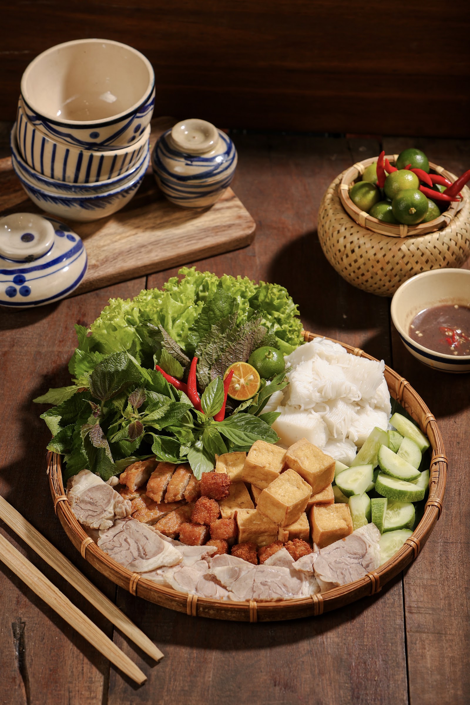 The bún đậu Quán Gánh platter on a round woven tray: golden fried tofu, sliced boiled pork belly, vermicelli, herbs, cucumber and a bowl of mắm tôm — stacked blue-white bowls and a basket of limes and chilies behind