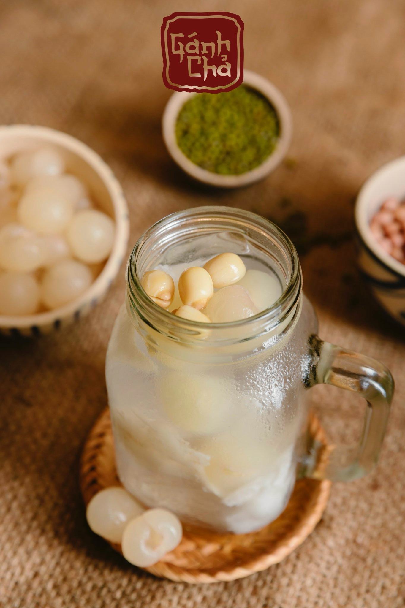 Chè sen nhãn nước dừa in a frosted mason mug on a woven coaster, with bowls of longan, mung beans and peanuts nearby — the Gánh Chả brand stamp on top