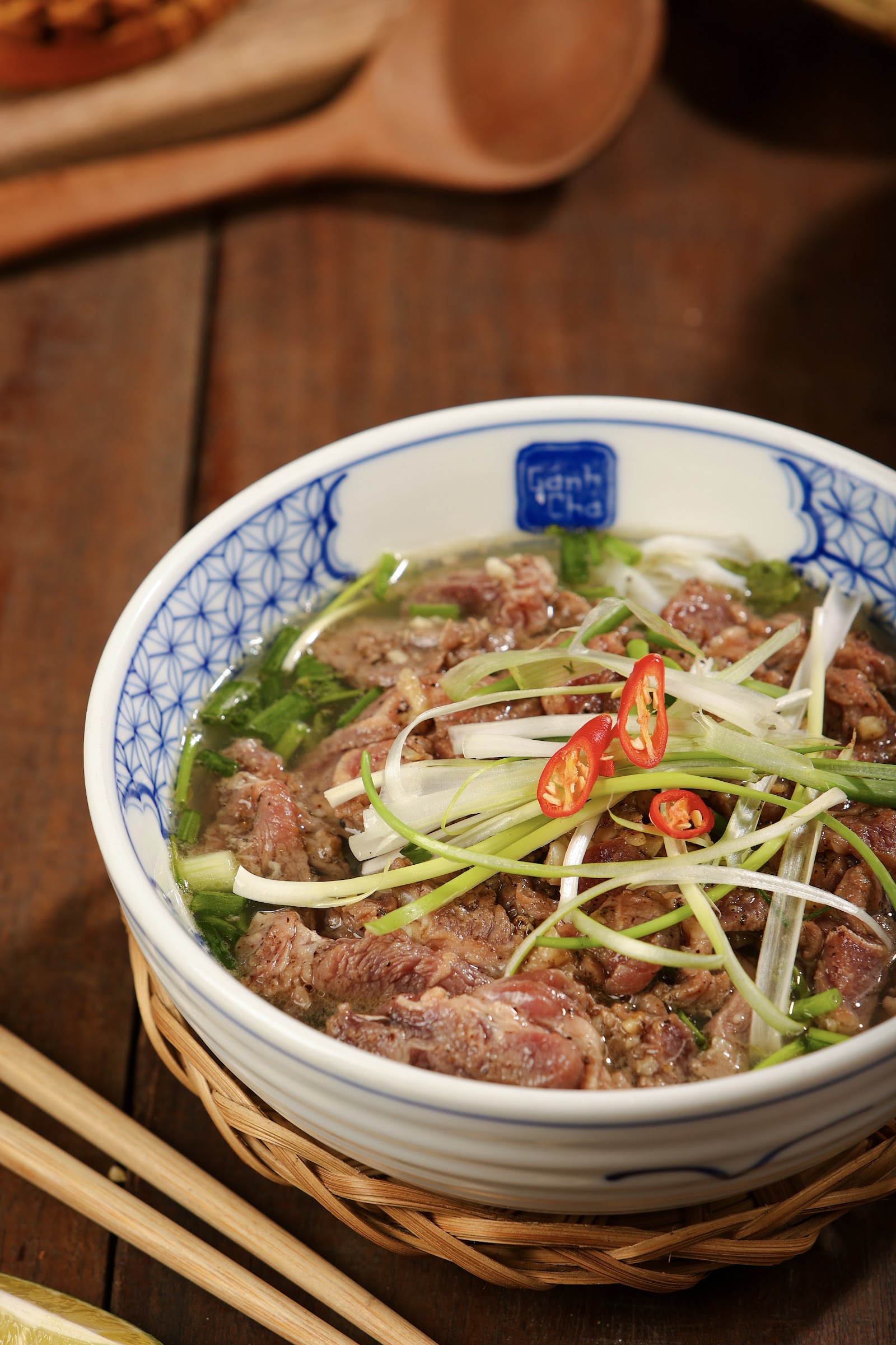 Phở bò tái lăn in the blue-white bowl stamped Gánh Chả, on a woven tray — seared beef, scallions, sliced chili