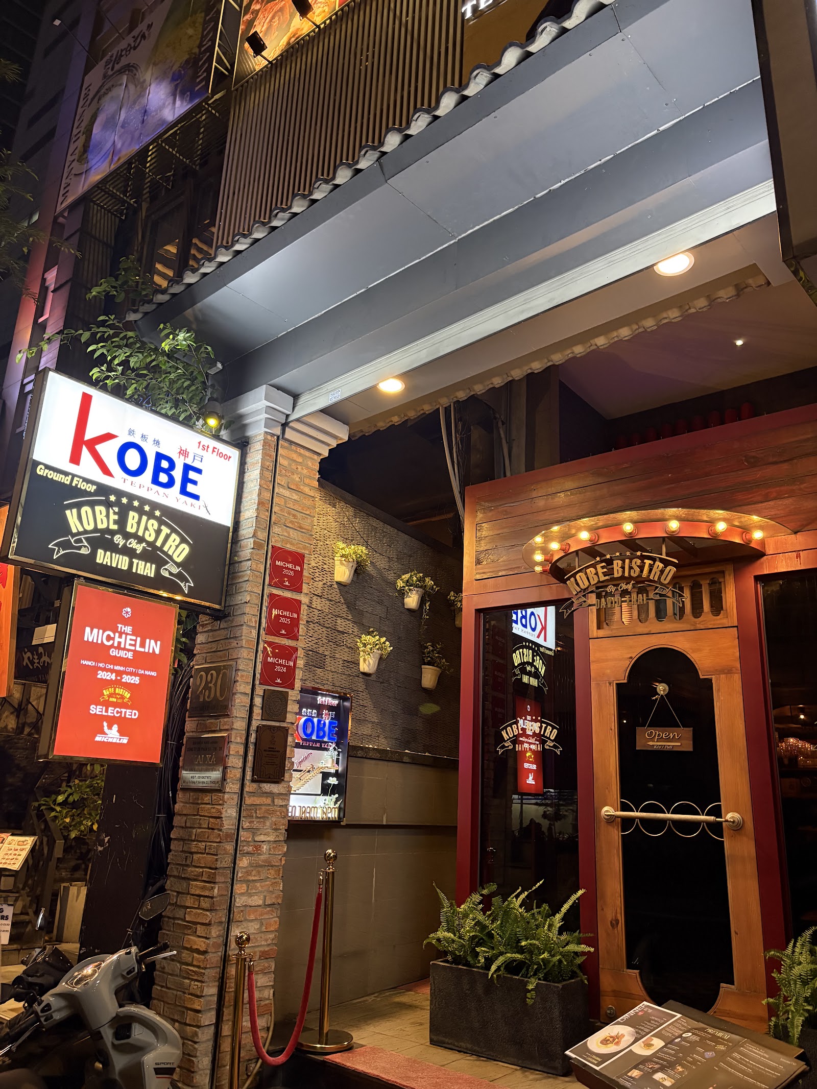 Kobe Bistro's entrance at night: lit signage, Michelin plaques, wooden door