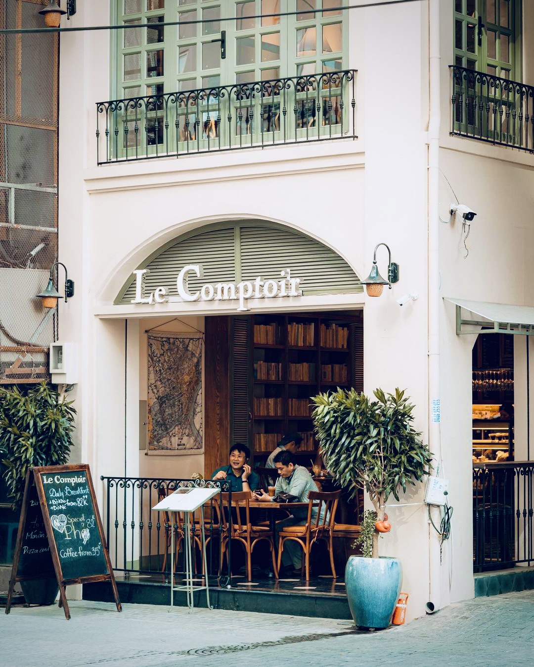 The Le Comptoir storefront at 12 Ho Huan Nghiep — the white slab-serif sign over the arched doorway, sage shutters, sidewalk tables with diners