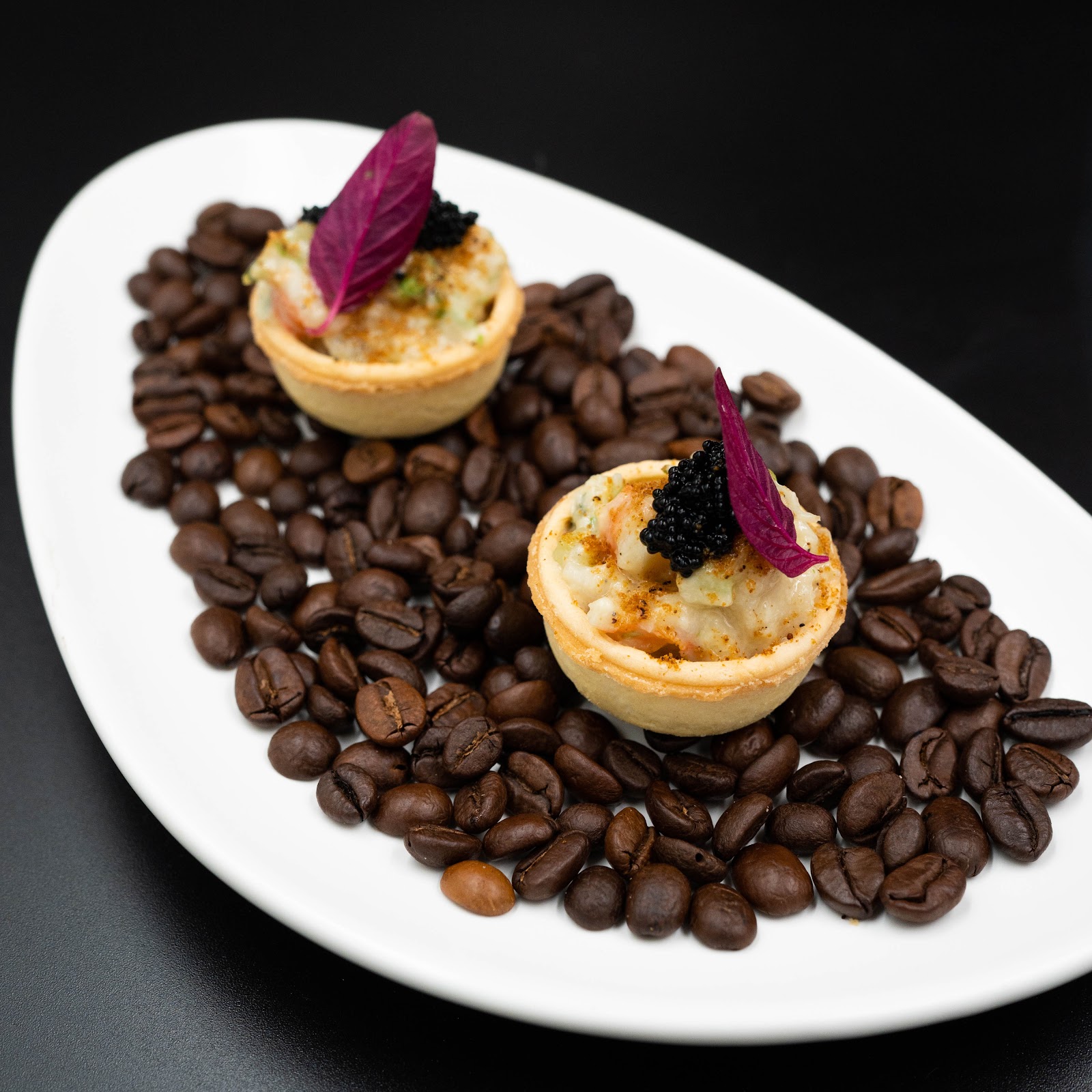 Two seafood tartlets topped with black caviar and a red shiso leaf, served on a white plate over coffee beans