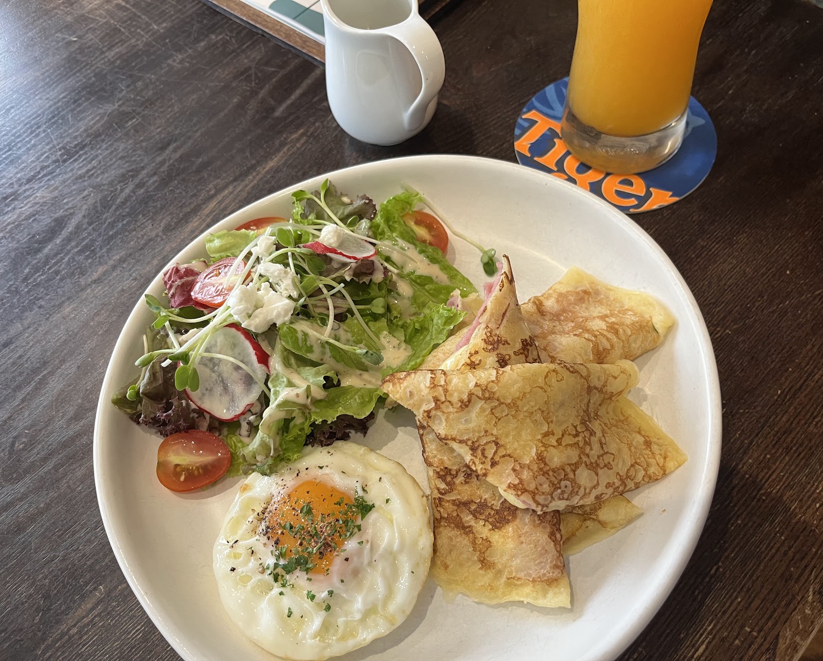 A plate of folded ham-and-cheese crêpes with a fried egg, a dressed green salad with radish and tomato, and orange juice on a dark wooden table