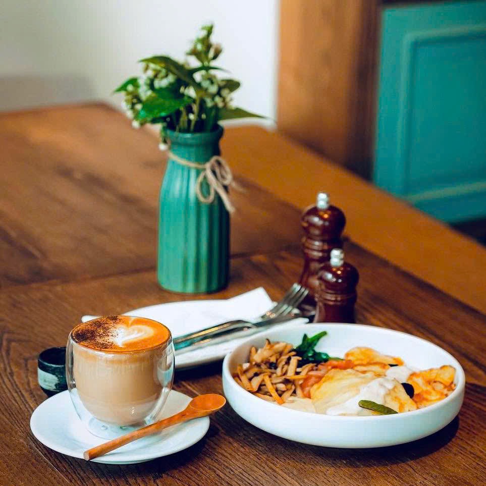 A glass-cup latte dusted with cocoa beside a white breakfast bowl of salmon, egg and mushrooms on a wooden table, a vase of flowers behind