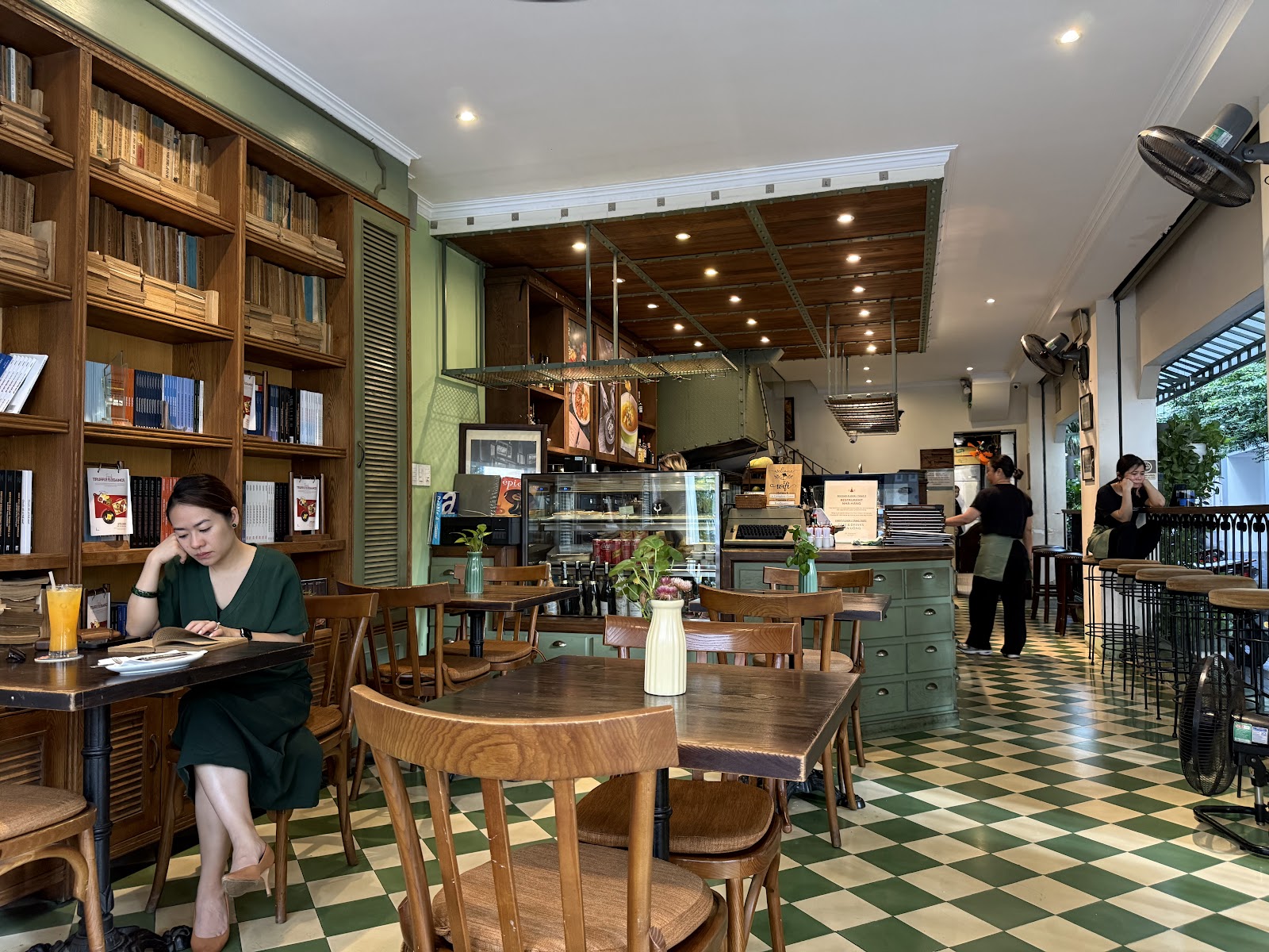 The library wall — floor-to-ceiling wooden bookshelves beside dark wood tables, a reader with coffee, the green-and-cream checkerboard floor