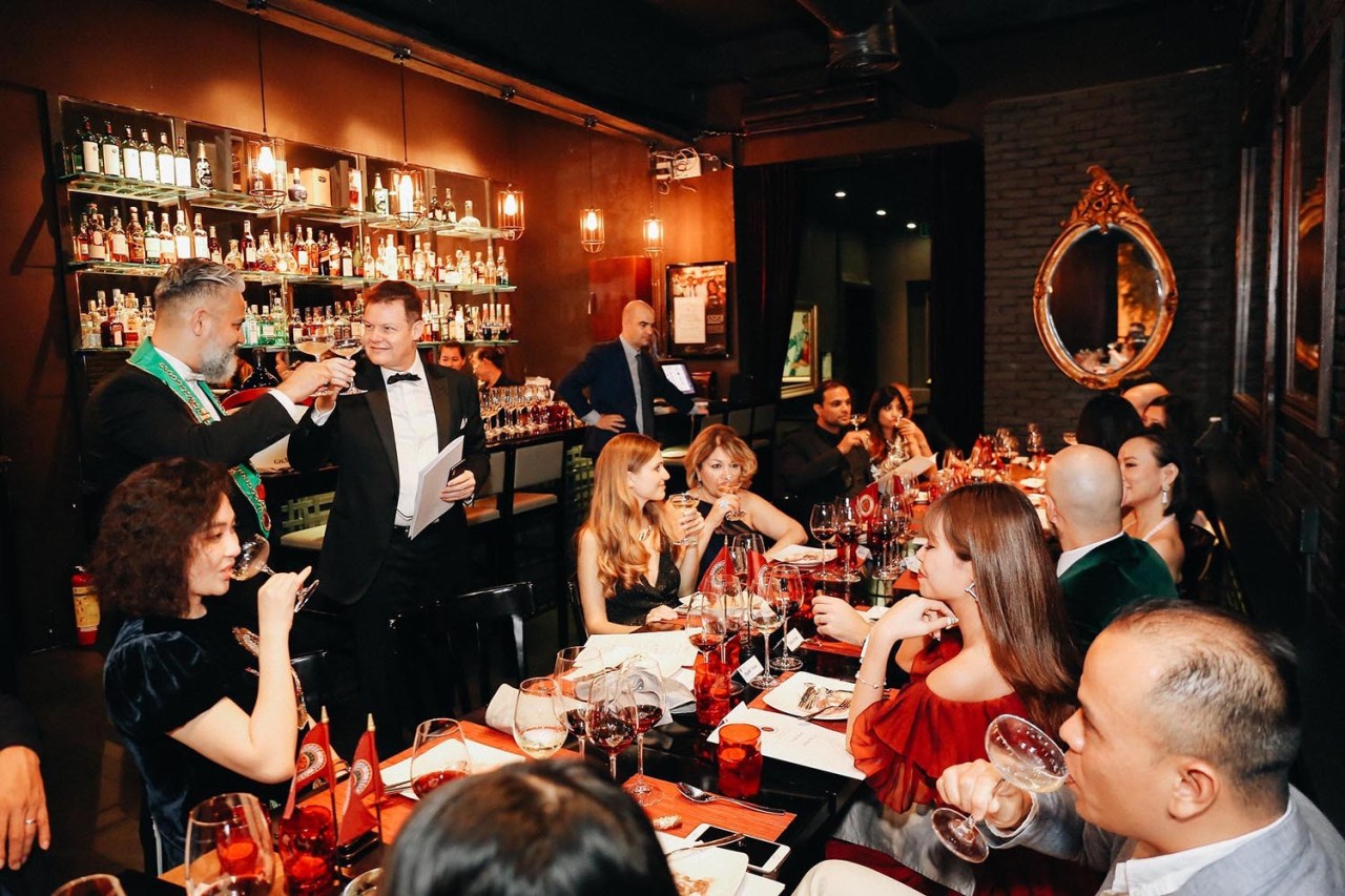 Gala dinner: guests toasting with wine glasses in the dark wood, candle-lit room with the bottle-lined back bar and a gilt mirror