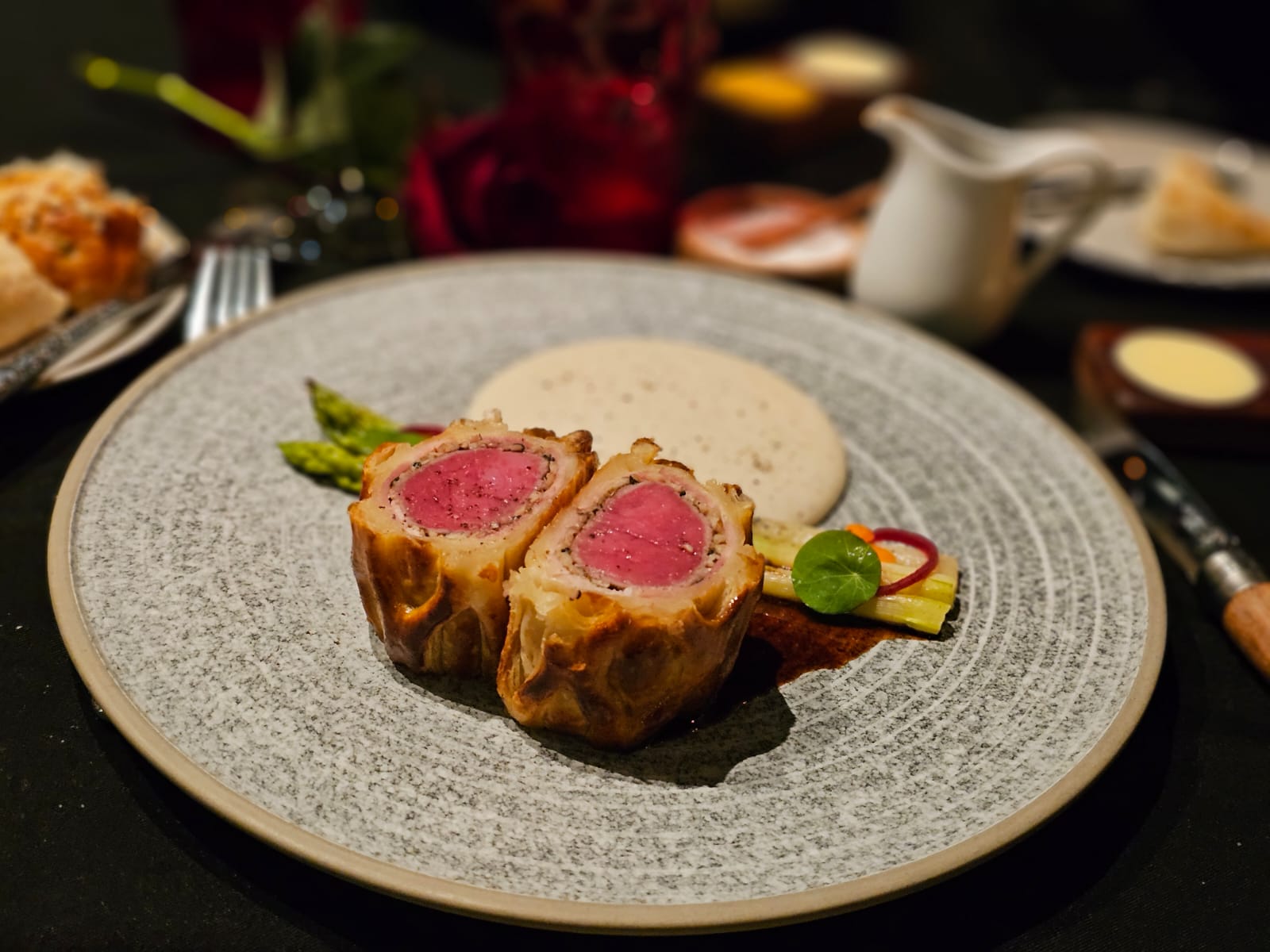 Two slices of beef wellington — pink-centered fillet, golden pastry — on the grey speckled plate; a white sauce jug, red wine glasses and candlelight at the table