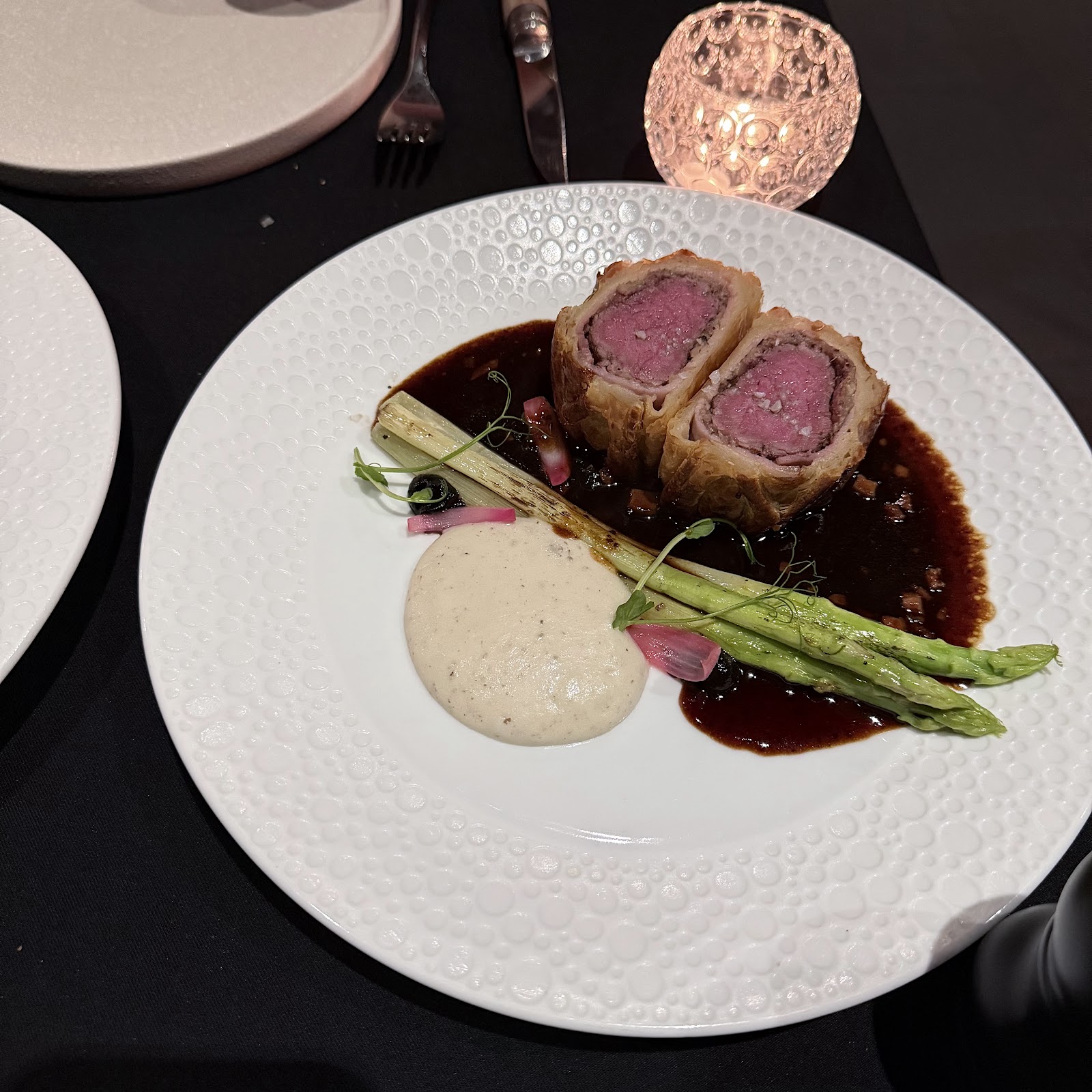Beef wellington on the white dotted plate with dark jus, asparagus and cream, a crystal candle glowing behind