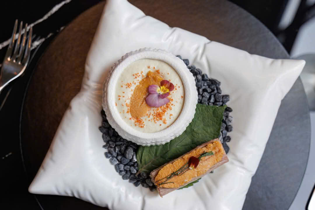 Overhead of sea urchin in a textured white shell bowl — orange uni, roe dust, a pansy — on a pillow-white porcelain plate with black pebbles, a shiso leaf, and a slice of pressed fish topped with uni