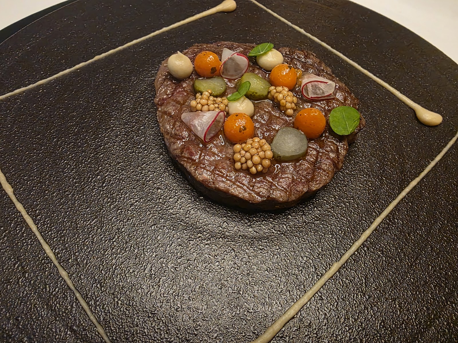 Finely chopped beef tartare on a dark textured plate — orange yolk spheres, mustard seeds, radish slices, herbs, and a square frame of sauce