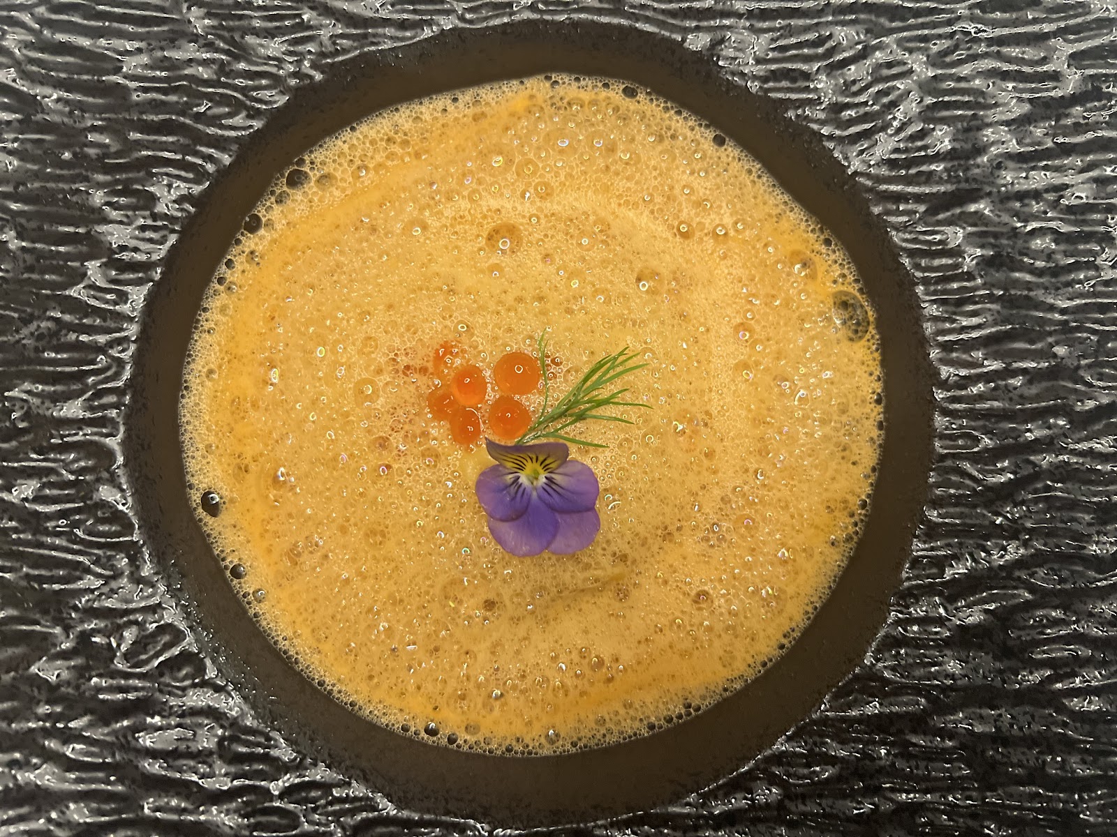 Overhead of frothy amber bisque in a dark rippled bowl — salmon roe, dill, a violet pansy