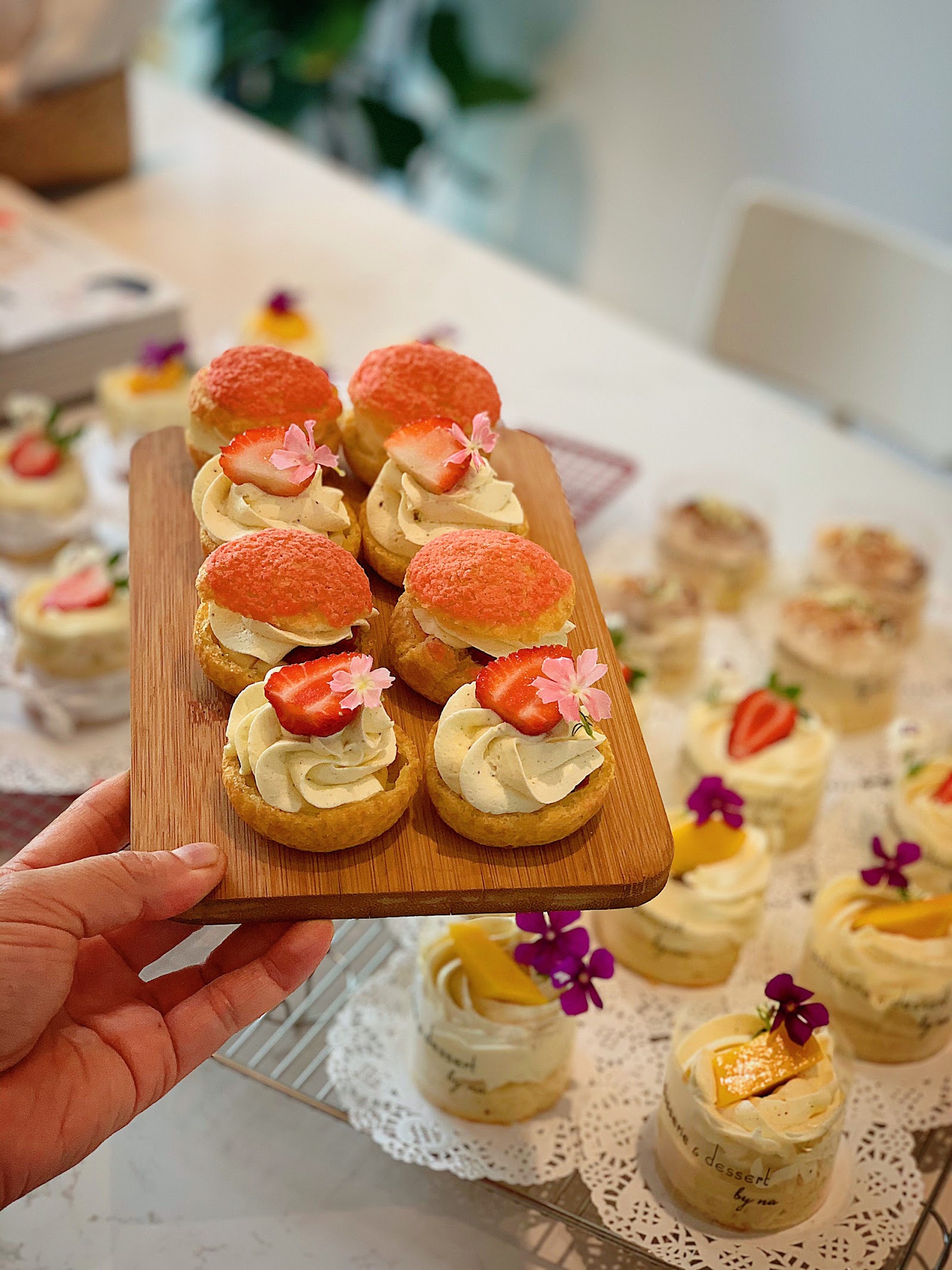 Choux pastries with strawberries and edible flowers on a wooden board — small flower-topped desserts behind
