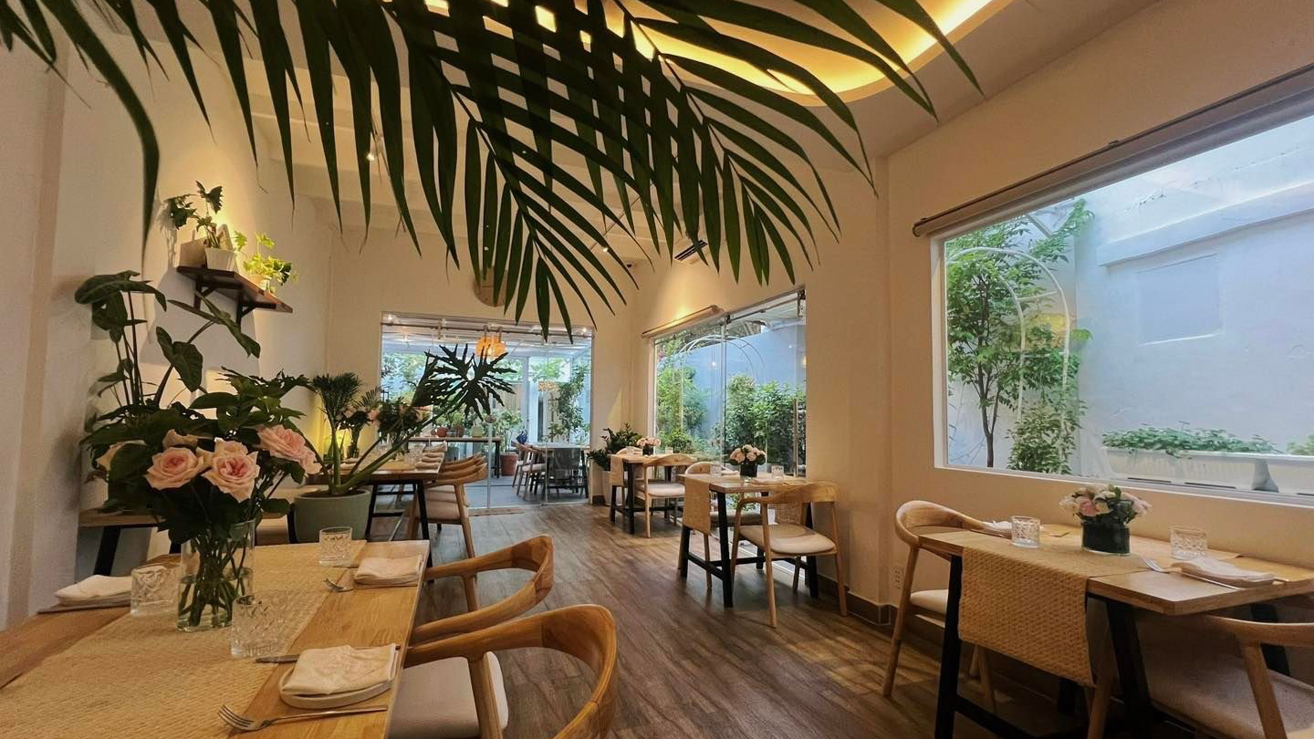 Maison des Rêves' plant-filled dining room — palm fronds, wooden tables, rattan chairs, a big garden window, roses on the table