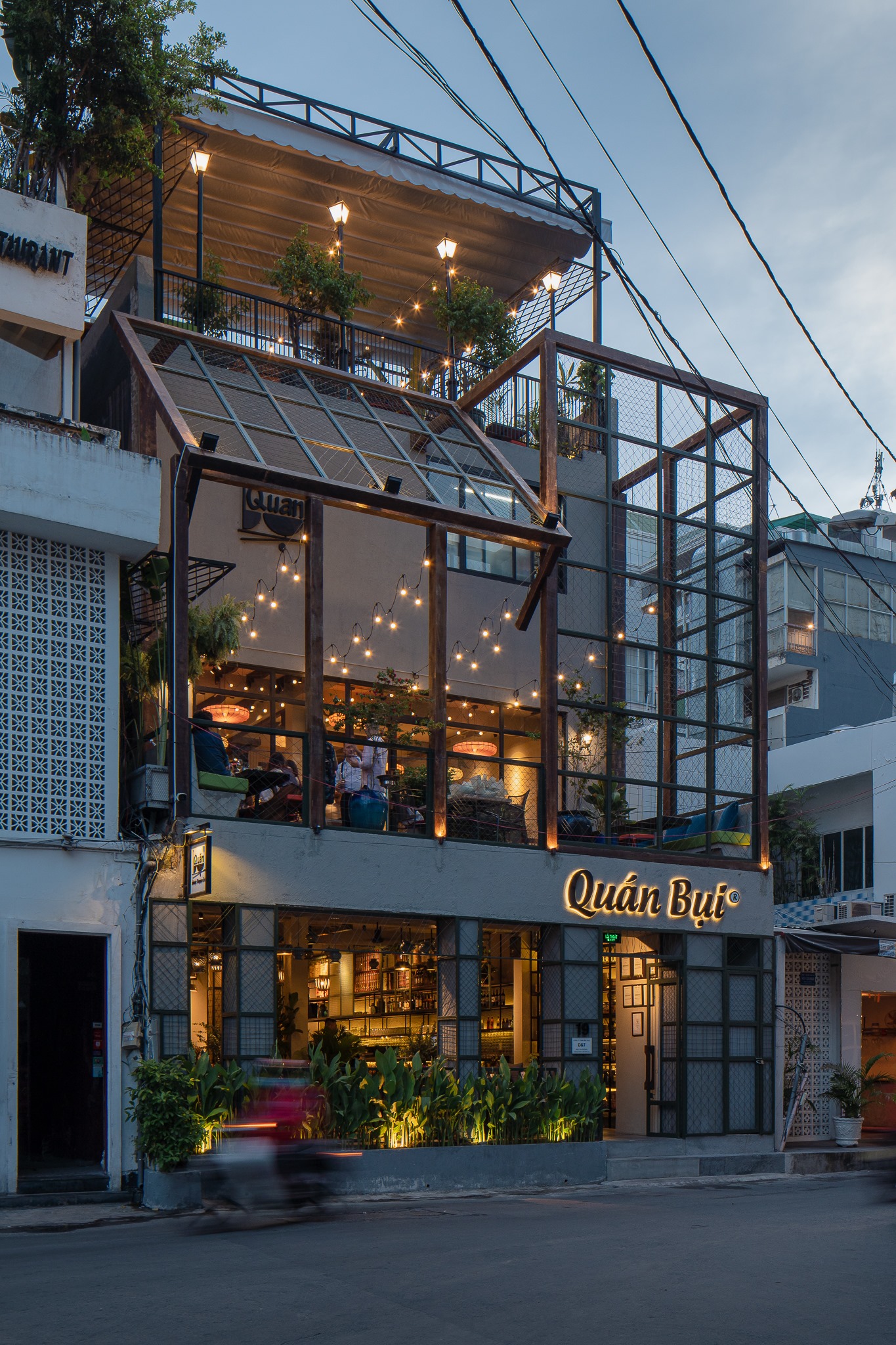 The Quán Bụi facade at dusk — glass frames, wooden architecture and the glowing sign