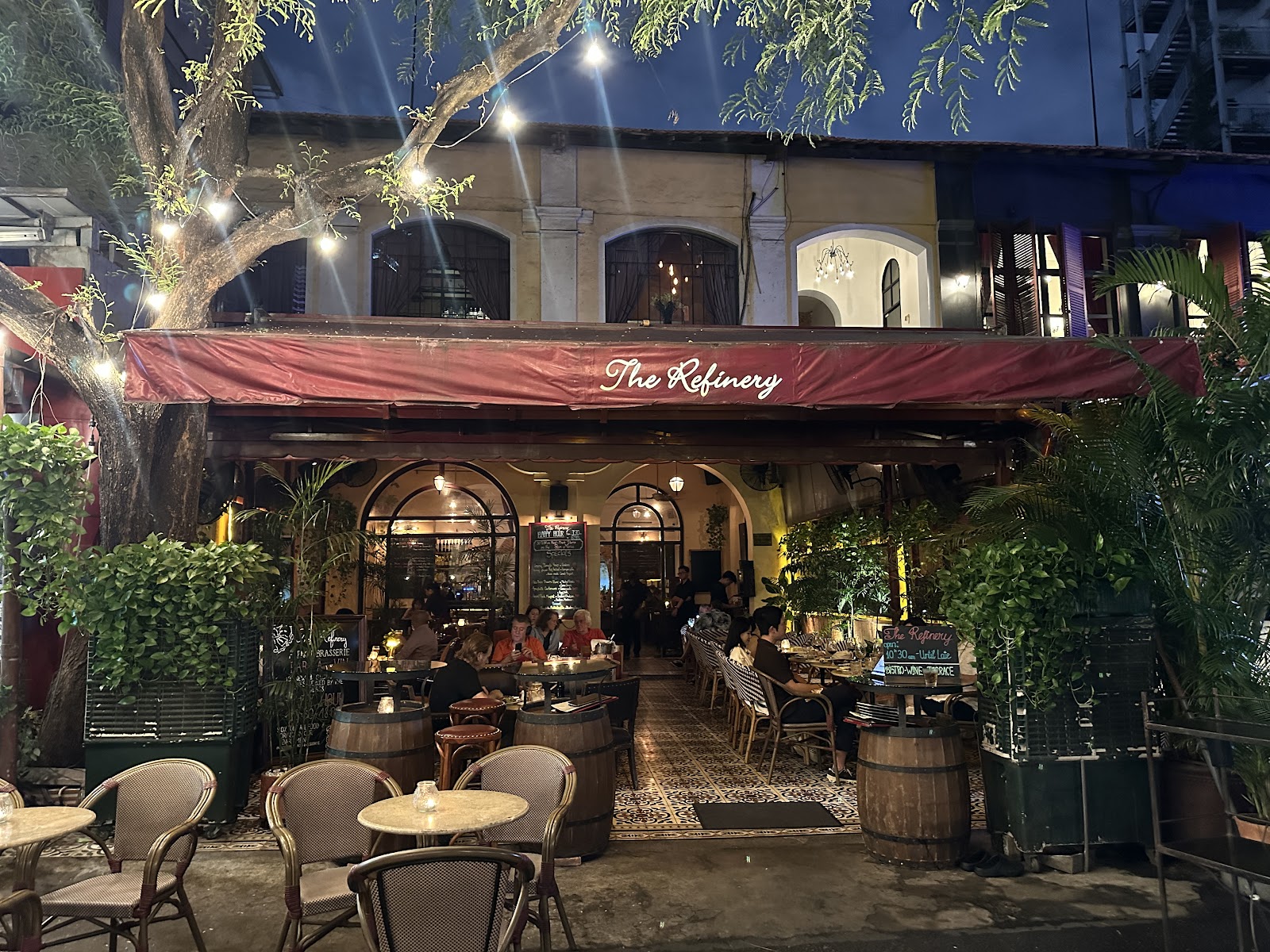 The courtyard at night with string lights in the big tree, the 'The Refinery' awning, diners around the barrel tables