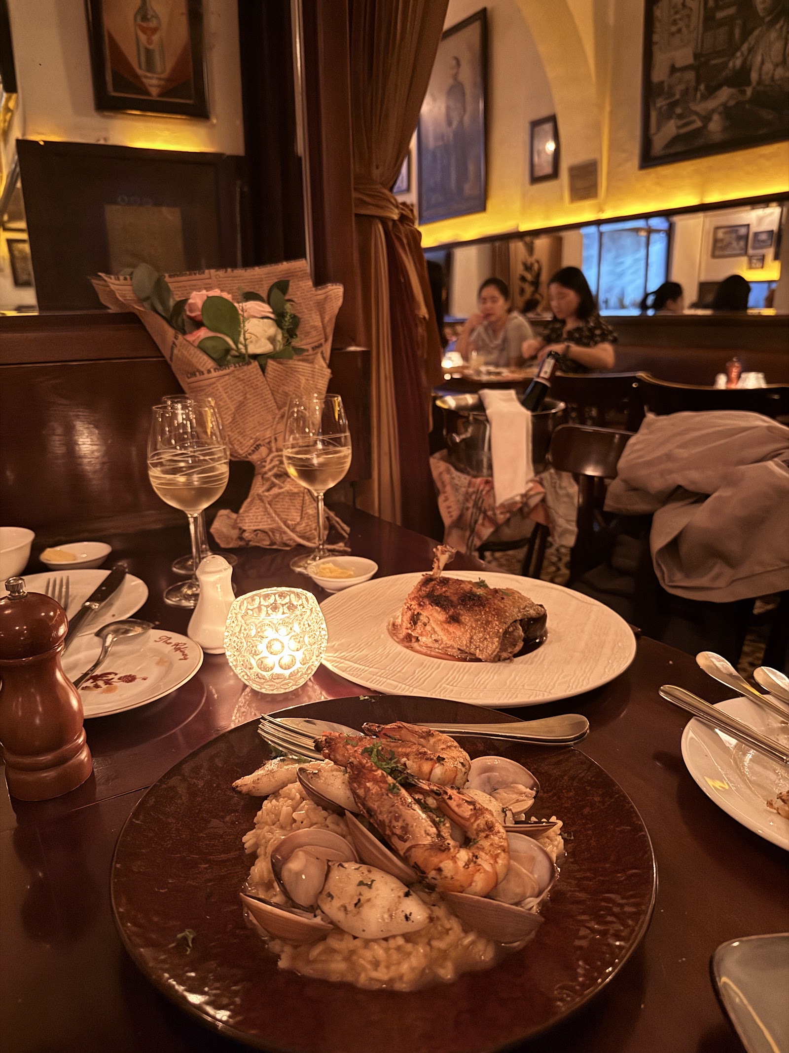 Seafood rice with grilled prawns and clams on a dark plate, with two glasses of white wine, a candle and flowers at the table — old-Saigon photos on the wall behind