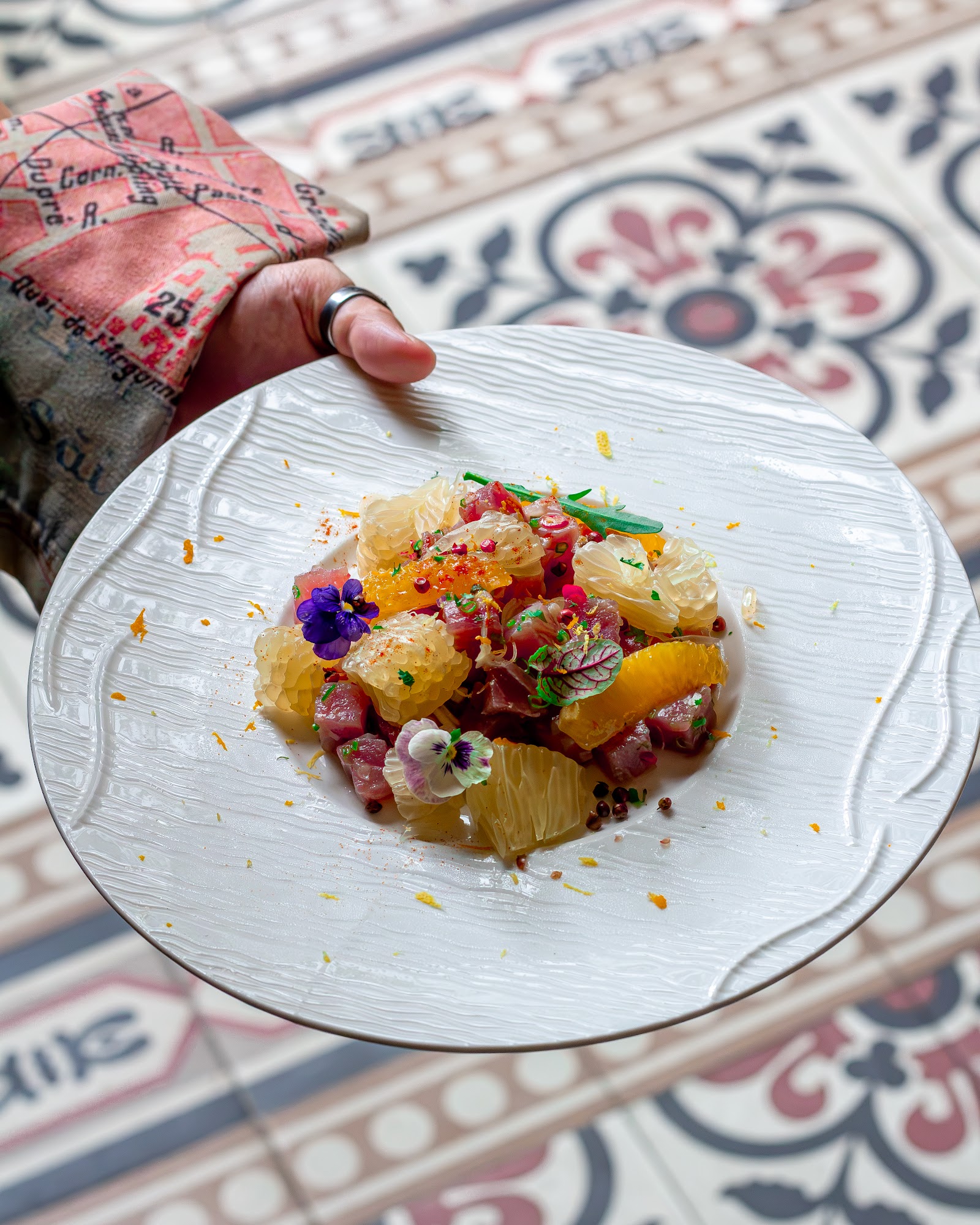 Fish tartare with pomelo and orange segments, purple and white edible flowers, herbs — on a white plate over patterned floral tiles