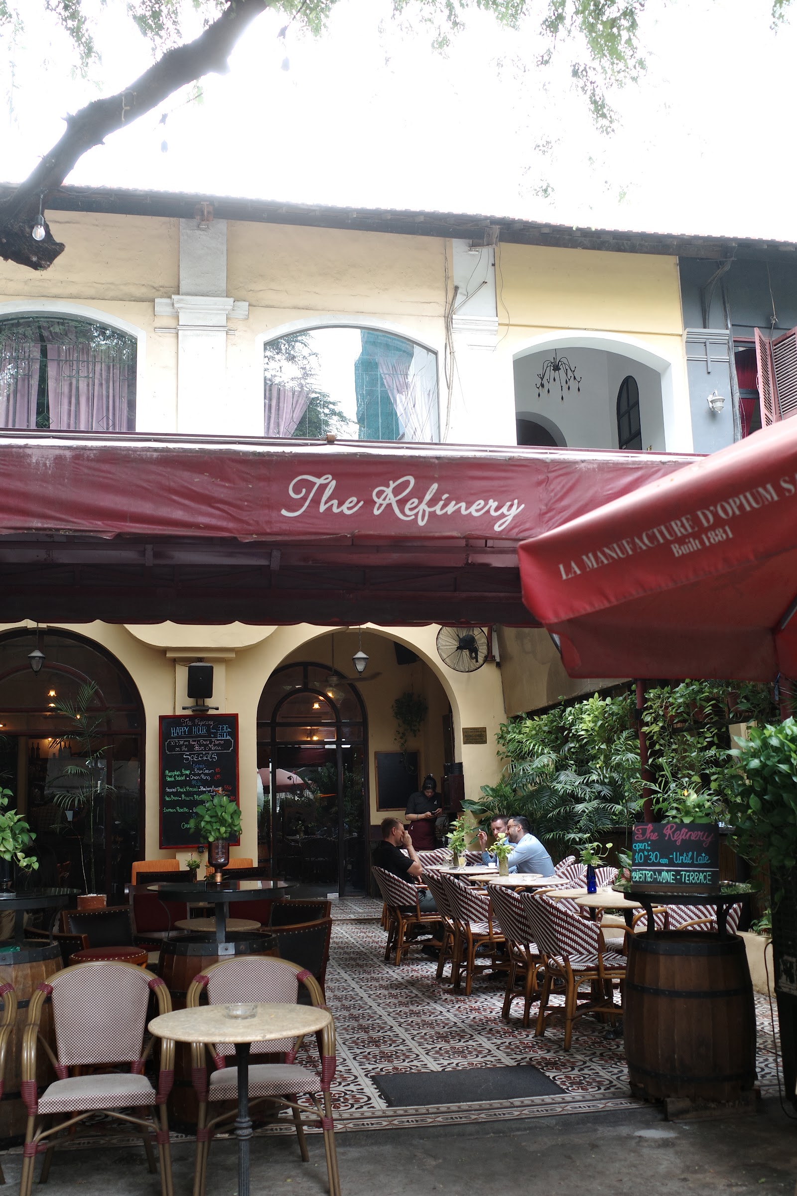 Looking up at the 'The Refinery' awning and the 'La Manufacture d'Opium Saigon — Built 1881' sign, diners on rattan chairs in the courtyard